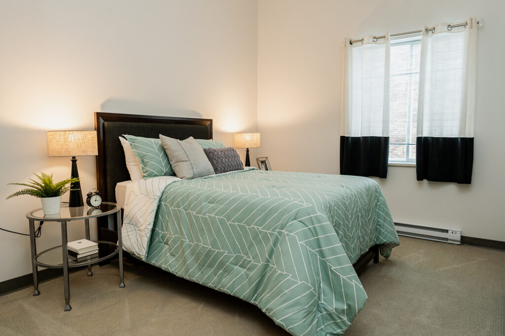 A made-up bed with light green sheets and gray throw pillows, a glass bedside table with a lamp, a potted plant, books, and an alarm clock on it.