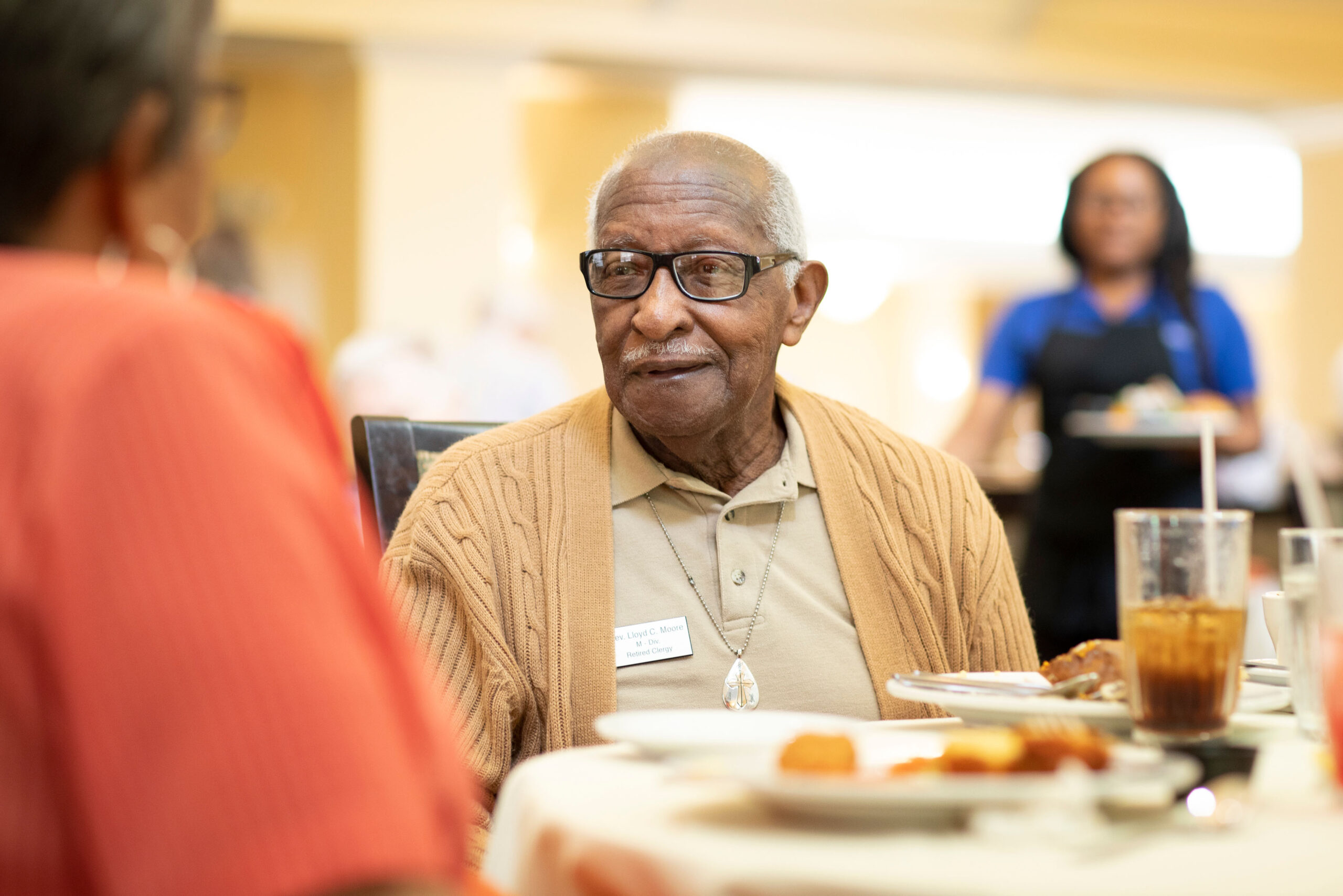 Grace_Management_Memphis_2018-28 - The Residence at Whitehall An older black man wearing classes and a beige cardigan is sitting at the dining table and conversing with another resident.