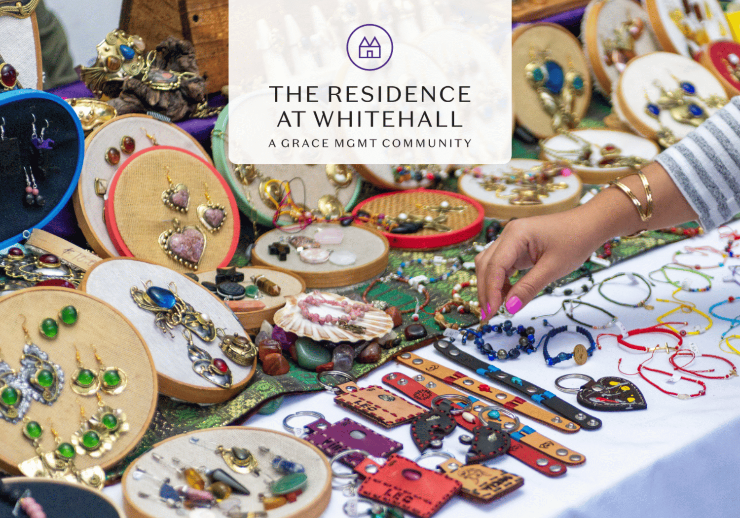 Colorful handmade jewelry spread across circular display boards at a market stall; a hand with pink nails reaches for bracelets under a banner reading 'The Residence at Whitehall'.