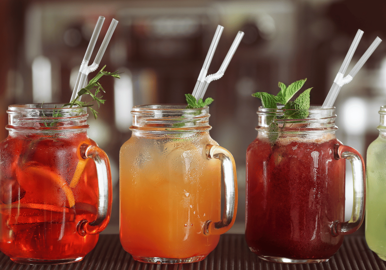 Three colorful fruit drinks in mason jars with mint garnish and white straws on a bar counter.