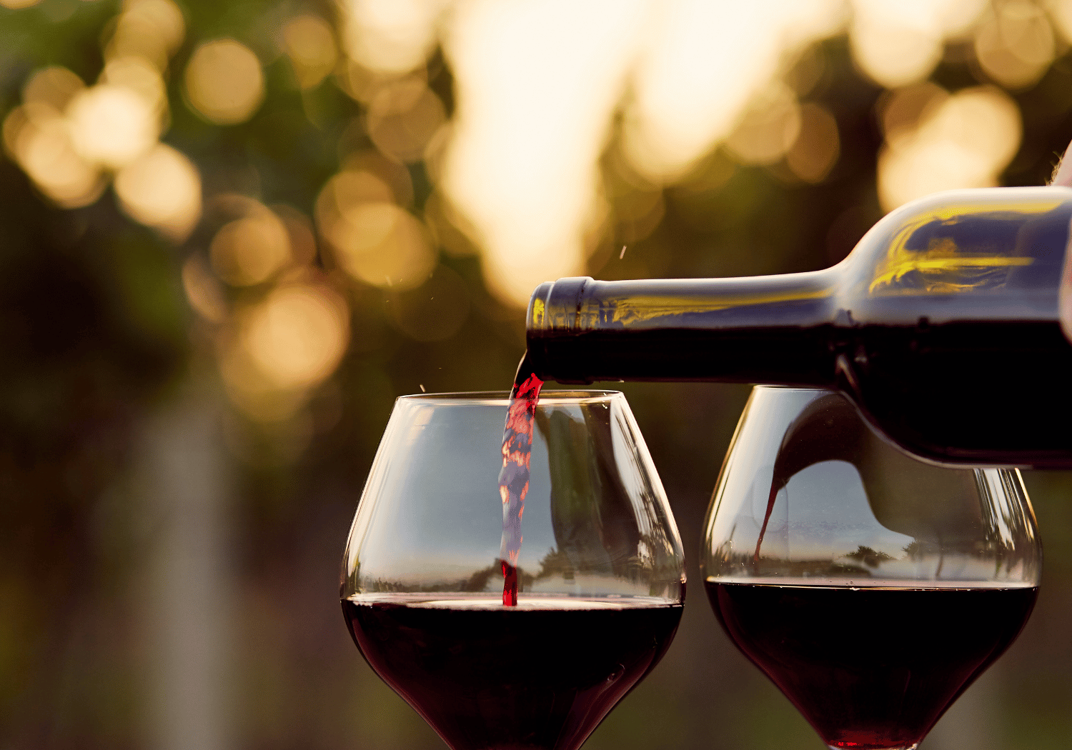 Red wine is being poured from a bottle into two wine glasses outdoors at sunset with a warm bokeh background.