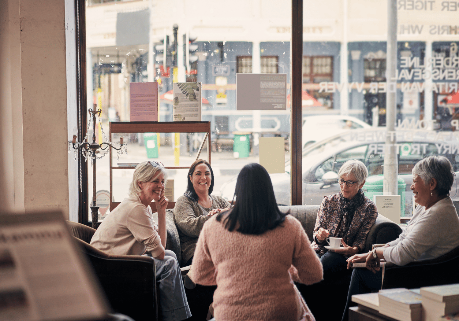 Five women sit together on a sofa in a cozy cafe, chatting and smiling by a large window.