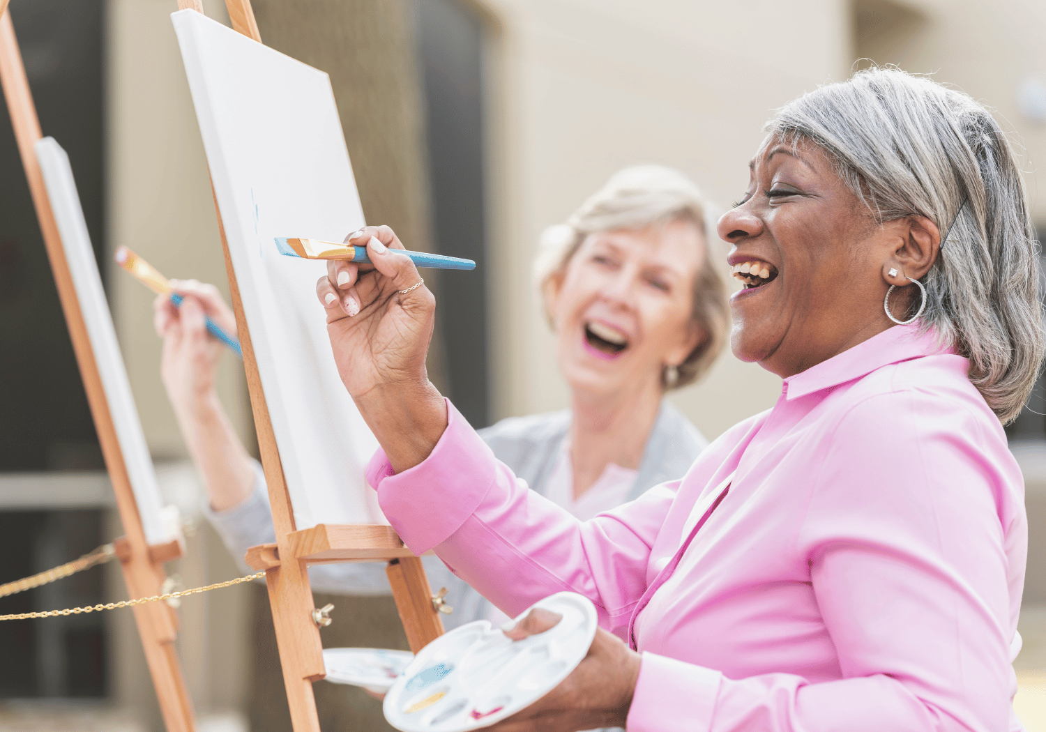 Two smiling women painting on easels outdoors, holding brushes and palettes.